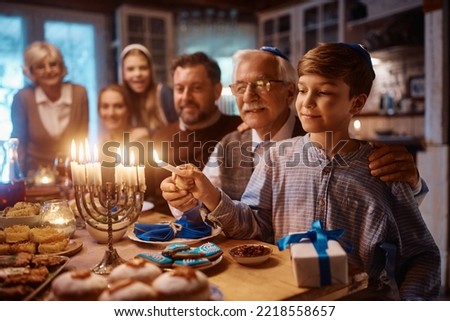 Happy extended Jewish family celebrating Hanukkah while gathering at dining table. Focus is on boy lighting candles in menorah. Royalty-Free Stock Photo #2218558657