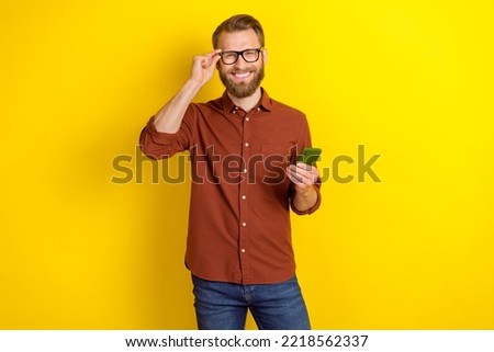 Photo of good mood cheerful man with blond beard dressed burgundy shirt hand touch glasses hold phone isolated on yellow color background
