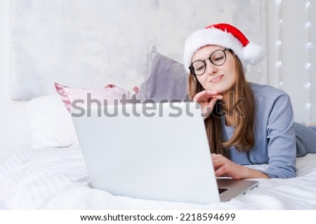 Happy girl in santa hat greets friends merry christmas video chat on laptop. Young woman lying on bed in blue pajamas with decorated pine tree in home interior with copy space