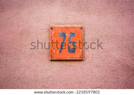 Plastic house number sevent five. Black lettering on a orange plate, mounted on a pinkish plastered wall