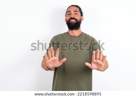 Serious Caucasian man with beard wearing green t-shirt over white background pulls palms towards camera, makes stop gesture, asks to control your emotions and not be nervous