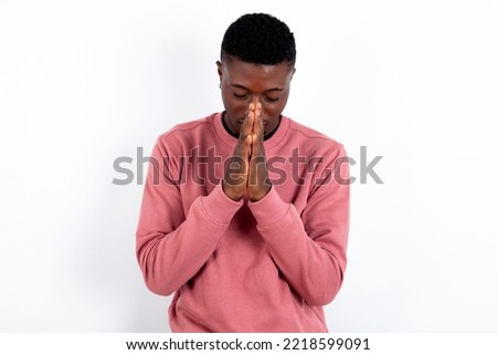 Indoor closeup of handsome man wearing pink sweater over white background practicing yoga and meditation, holding palms together in namaste, looking calm, relaxed and peaceful.