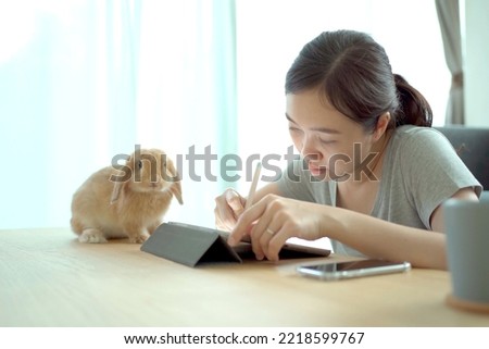 Happy cheerful Asian woman enjoy drawing the illustration on digital tablet and playing with her pet, a lovely Holland Lop bunny on the wooden table. Woman drawing or sketching bunny picture on tablet