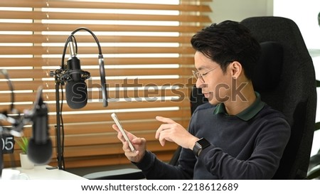 Young asian man radio host sitting front of condenser microphone and using mobile phone