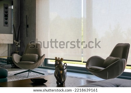 Interior design with roller blinds in the background. Automatic solar shades of large sizes on the window. Fabric with linen texture. In front of a large window is a chair on a carpet.  Royalty-Free Stock Photo #2218639161