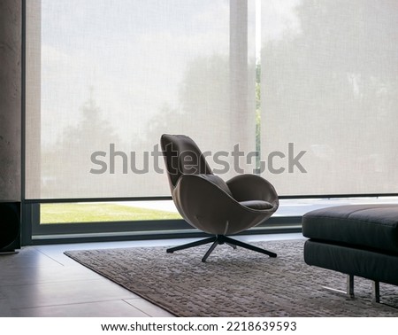 Roller blinds of large sizes on the window in the interior. Automatic solar shades, fabric with linen texture. In front of a large window is a chair on a carpet. Outside is a view of the garden. Royalty-Free Stock Photo #2218639593