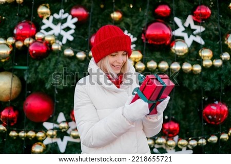 A woman in a knitted red hat with a surprised look holds a gift box in her hands while standing at a Christmas tree in the city square. The girl is astonished by the present