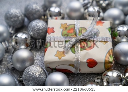 shiny gift box with christmas, new year print decorations and tied bow, surrounded by silver balls baubles on grey sparkle background, festive holidays time