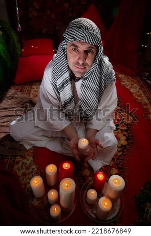Arab sheikh in a white robe and a headscarf in a boudoir in a room with a red carpet and candles with smoke. Oriental eastern fabulous style photo shoot with a man. Male model posing in fary tale