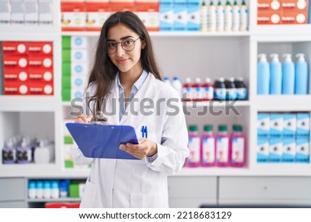 Young hispanic girl pharmacist writing on report clipboard at pharmacy