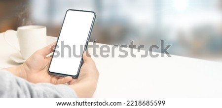 Mockup image of hand holding white mobile cellphone with blank screen with coffee cup on white table. wide banner mock up smartphone