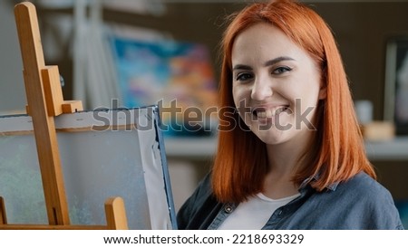 Caucasian woman artist girl painter with red hair enjoying art work with paints creates picture drawing painting on canvas looking at camera smiling posing smile toothy happy cheerful draw laughing