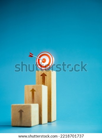 Target icon on top of wooden blocks with rise up arrows, 3d bar graph chart steps on blue background, vertical style, business growth process, profit, investment, economic improvement concepts. Royalty-Free Stock Photo #2218701737