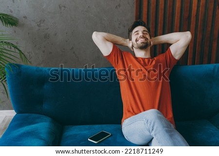 Young man wears red t-shirt listen music in earphones sit on blue sofa near mobile cell phone stay at home flat rest relax spend free spare time in living room indoors grey wall. People lounge concept