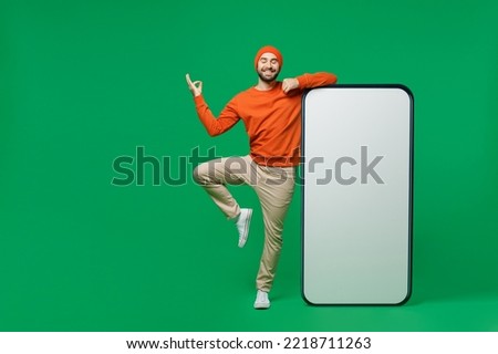 Full body young calm man 20s wear orange sweatshirt hat stand near big blank screen mobile cell phone with workspace copy space mockup area do yoga om gesture isolated on plain green background studio