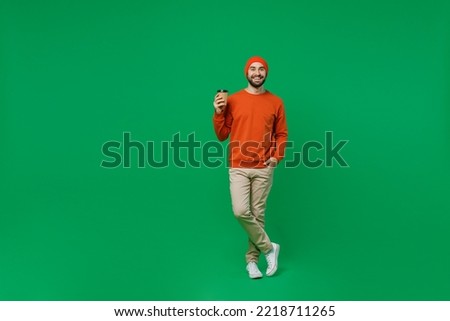 Full body young smiling happy man 20s wear orange sweatshirt hat hold takeaway delivery craft paper brown cup coffee to go isolated on plain green background studio portrait. People lifestyle concept