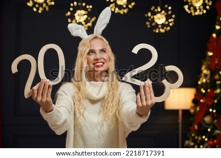 Front view of beautiful woman wearing knitted sweater, looking up, holding 2023 decoration, smiling. Attractive blonde girl with red lips standing, dreaming. Concept of new year and xmas.