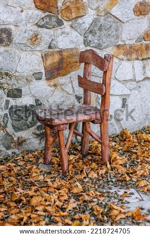 A large homemade vintage brown wooden chair stands on a park with yellow maple leaves in autumn against a stone wall.