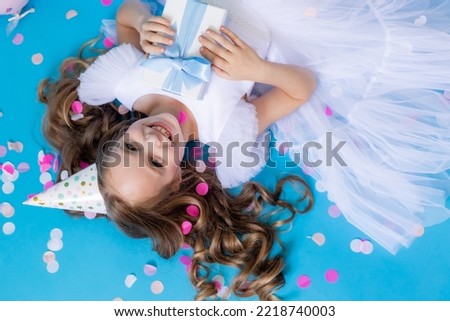Cute girl in a fluffy white dress with a gift in her hands lies on a blue background in confetti. High quality photo