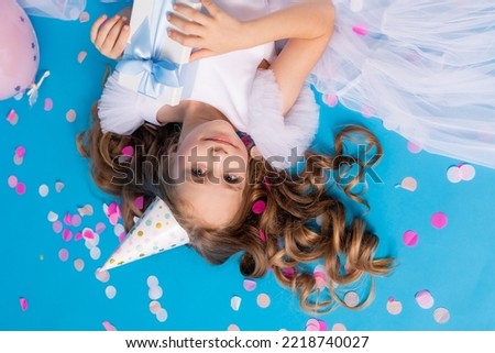 Cute girl in a fluffy white dress with a gift in her hands lies on a blue background in confetti. High quality photo