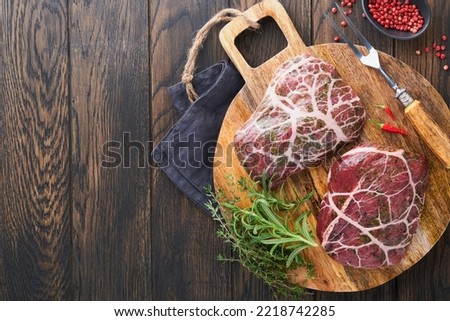 Steaks Raw. Raw cowboy steak, marbled beef meat with spices rosemary and pepper on black wooden board on old wooden dark table background. Top view. Mock up.