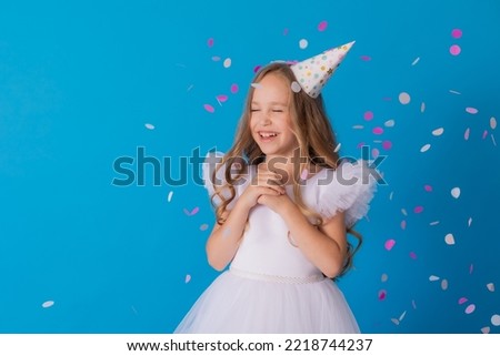 girl in a beautiful dress and a cardboard hat stands on a blue background and throws confetti. High quality photo