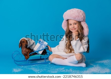 studio photo of a girl in a white sweater and hat and a dachshund dog sitting in a sled. High quality photo
