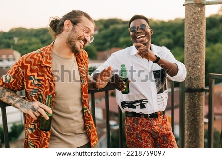 Two multiracial friends have fun at the open-air summer rooftop party, tell jokes and laugh while holding a beer and leaning on the railing. Royalty-Free Stock Photo #2218759097