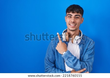 Young hispanic man standing over blue background with a big smile on face, pointing with hand finger to the side looking at the camera. 