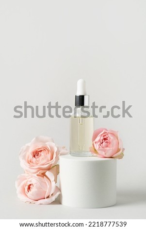 Serum in transparent glass dropper bottle on white round podium and pink roses on light grey background. Natural skin care product. Beauty routine still life. Trendy product for branding. Front view.