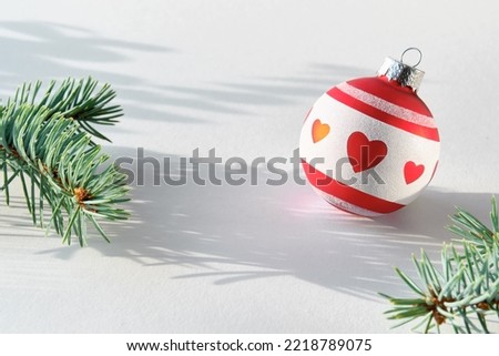 Single Christmas decorative trinket. Red white glass ball with heart shapes. Fir twig with frosted cone. Sunlight with long shadows, off white background. Festive Xmas design.