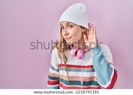 Young blonde woman standing over pink background smiling with hand over ear listening an hearing to rumor or gossip. deafness concept. 