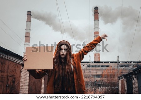 One woman holds a cardboard poster in her hands against standing background of the factory. Activist in orange hoodie during a picket with placard at the plant. Concept of environmental pollution.
