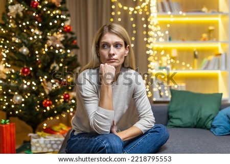 sad Woman alone at home for Christmas and New Year, sitting depressed near Christmas tree on sofa in living room, waiting for celebration.