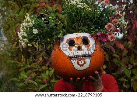 Woman Holds The Big Pumpkin With Chrysanthemum Flowers. Festive Decoration For Day Of The Dead In The Autumn Garden. Día de Muertos Background. Funny Skull (Calavera de Azúcar)