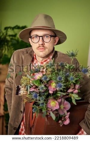 Poet or writer portrait. Nerd concept: caucasian male person in eyeglasses and hat staying with flower bouquet and looking in camera in flower store. High quality vertical image
