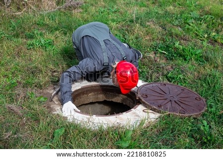 A male plumber opened the hatch of a water well and looks inside. Inspection of water pipes and meters. Royalty-Free Stock Photo #2218810825