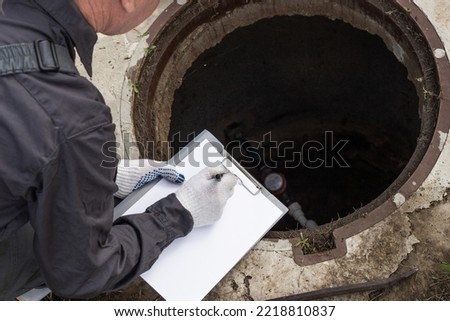 A male plumber near a water well records the measurements taken and the readings of the water meter. Royalty-Free Stock Photo #2218810837