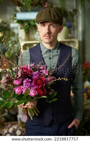 Women's Day or Valentine's Day concept. Attractive male in peaked cap and vintage clothes from 20s holding beautiful bouquet of red and violet flowers. Congratulating with flowers. High quality image