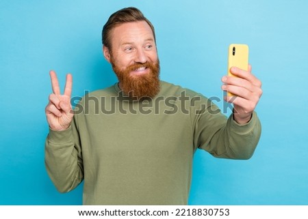 Photo of optimistic handsome guy with ginger hairstyle khaki long sleeve hold smartphone showing v-sign isolated on teal color background