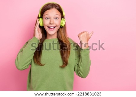 Photo of shiny impressed schoolgirl wear green sweatshirt earphones pointing thumb empty space isolated pink color background