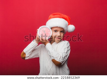 A boy in a Santa Claus hat holds a watch on a red studio background. the concept of Christmas and new year. a child in knitted clothes.