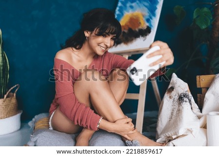Carefree young female artist posing for smartphone camera selfie near easel and paints showing in blog her hobby. Creative smiling brunette woman photographing herself near draw oil picture 