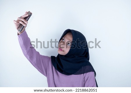 Excited joyful young brunette asian woman wearing hijab standing doing selfie shot on mobile phone waving greeting with hand isolated on white background, studio portrait