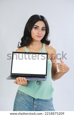 beautiful, young and intelligent-looking Pakistani Asian woman student wearing a sleeveless green top, holding a laptop looking in camera. Girl showing empty or blank laptop screen copy space