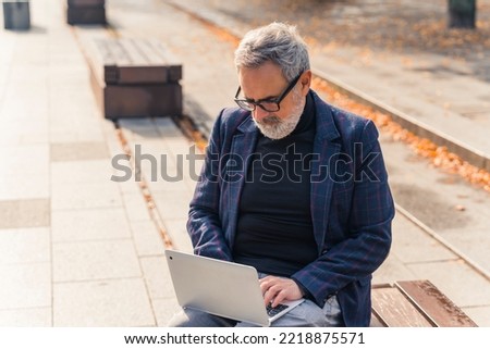 Modern stylish bearded grey-haired mature man entrepreneur sitting on a bench in the park with fall foliage and working on his laptop. High quality photo