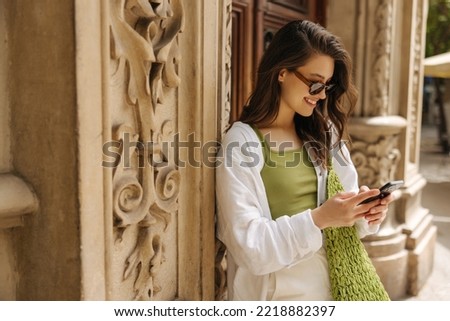 Smiling young caucasian woman reads pleasant sms on smartphone outdoors. Brunette wears casual clothes, sunglasses, bag. Good mood concept