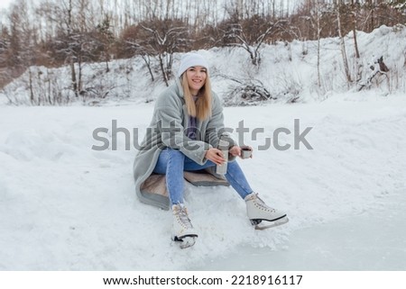 Lovely young woman relaxing after riding ice skates and drinking hot drink from thermo pot on the ice rink. Girl sitting on snow and smiling.