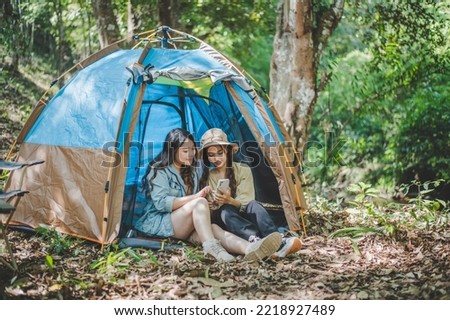 front view Young Asian pretty woman and her girlfriend sitting at front of tent, use mobile phone take photo during camping in forest with happiness together