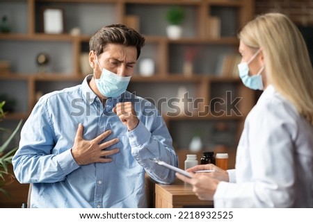 Coronavirus Concept. Sick Young Man Wearing Medical Mask Coughing At Appointment With Female Doctor, Ill Male Patient Suffering Respiratory Disease, Having Acute Bronchitis Or Pneumonia Royalty-Free Stock Photo #2218932525